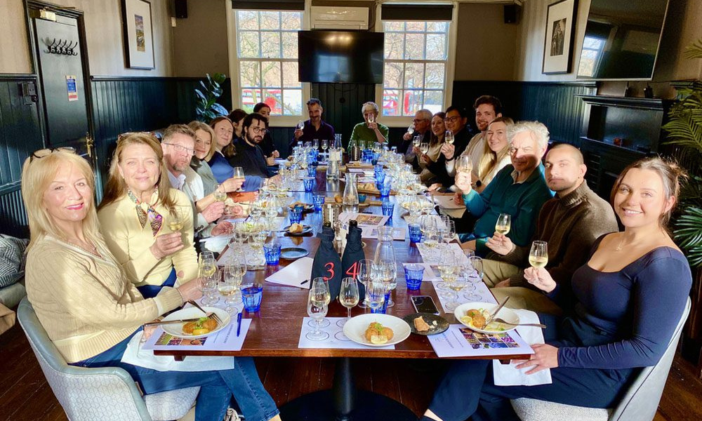 a table of 18 guests toasting the camera with tasting mats in front of them on the table