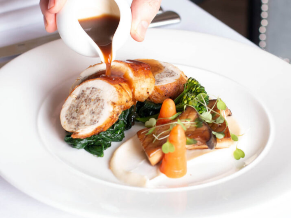 A smooth gravy being poured over a rolled chicken dishes plated with vegetables